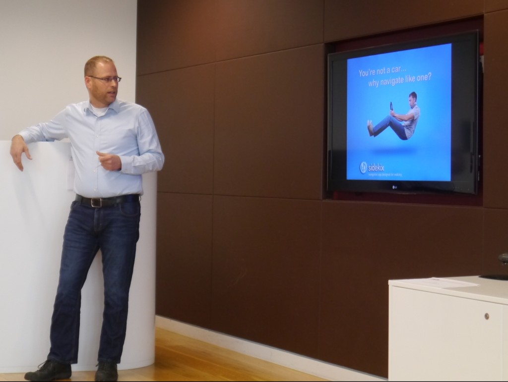 Sidekix co-founder Miron Perel stands next to a presentation screen. On it is a picture of a person floating in a sitting position, as if driving a car without the vehicle around him. The text reads, "You're not a car... why navigate like one?"
